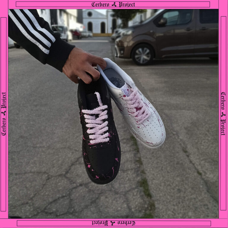 Person holding one black and one white sneaker, both featuring pink laces and paint splatter details
