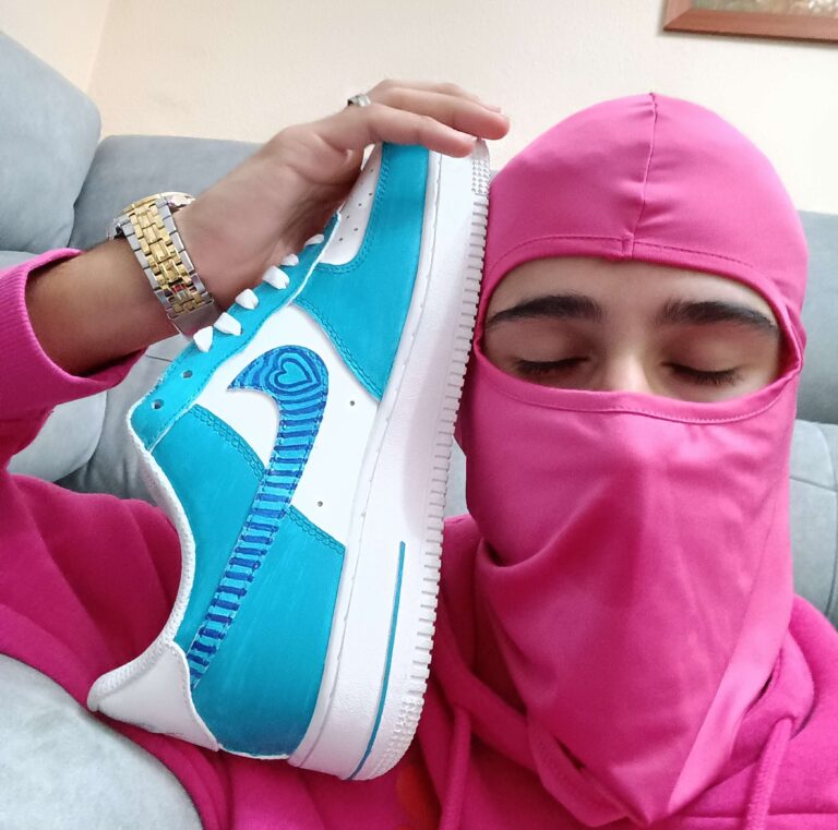 Person in pink balaclava holding a custom white and blue sneaker with a heart-shaped swoosh
